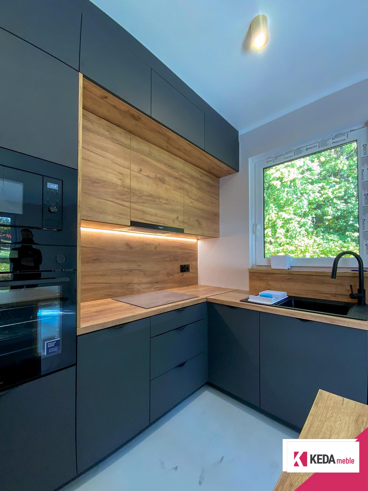 Nowoczesna kuchnia z szarymi szafkami i drewnianymi blatami, wbudowanymi urządzeniami AGD, dużym oknem z widokiem na zieleń oraz centralnie umieszczoną wyspą kuchenną.