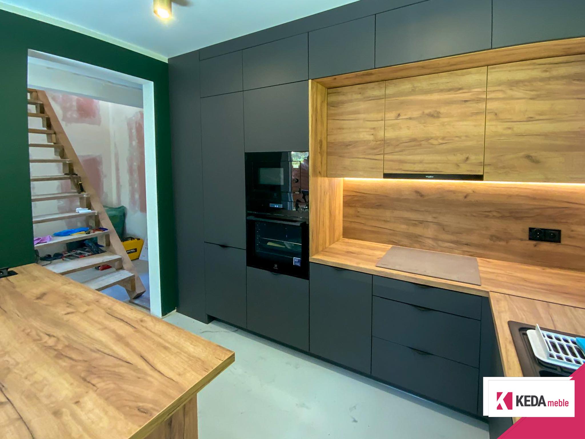 Nowoczesna kuchnia z szarymi szafkami i drewnianymi blatami, wbudowanymi urządzeniami AGD, dużym oknem z widokiem na zieleń oraz centralnie umieszczoną wyspą kuchenną.