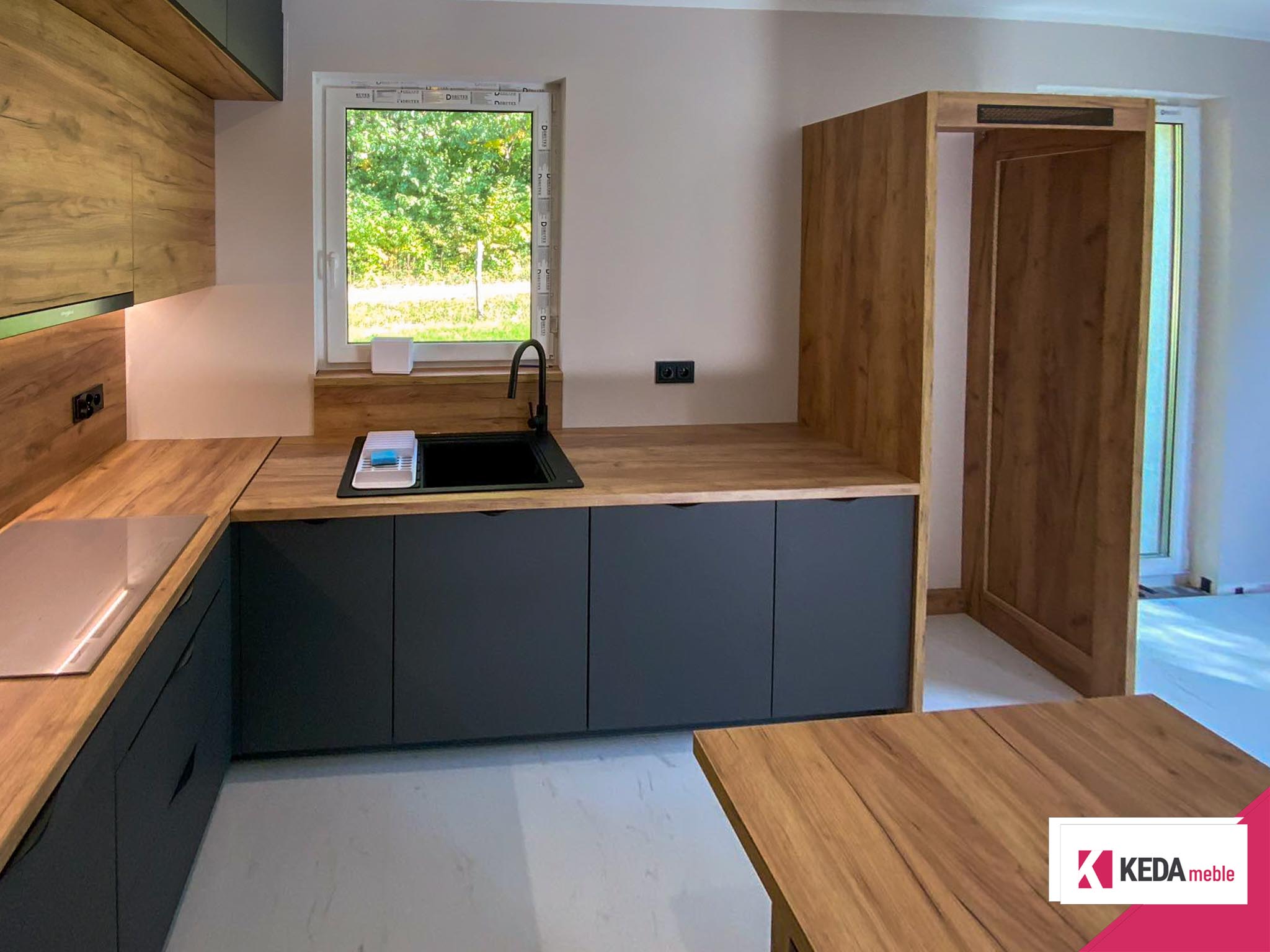 Nowoczesna kuchnia z szarymi szafkami i drewnianymi blatami, wbudowanymi urządzeniami AGD, dużym oknem z widokiem na zieleń oraz centralnie umieszczoną wyspą kuchenną.