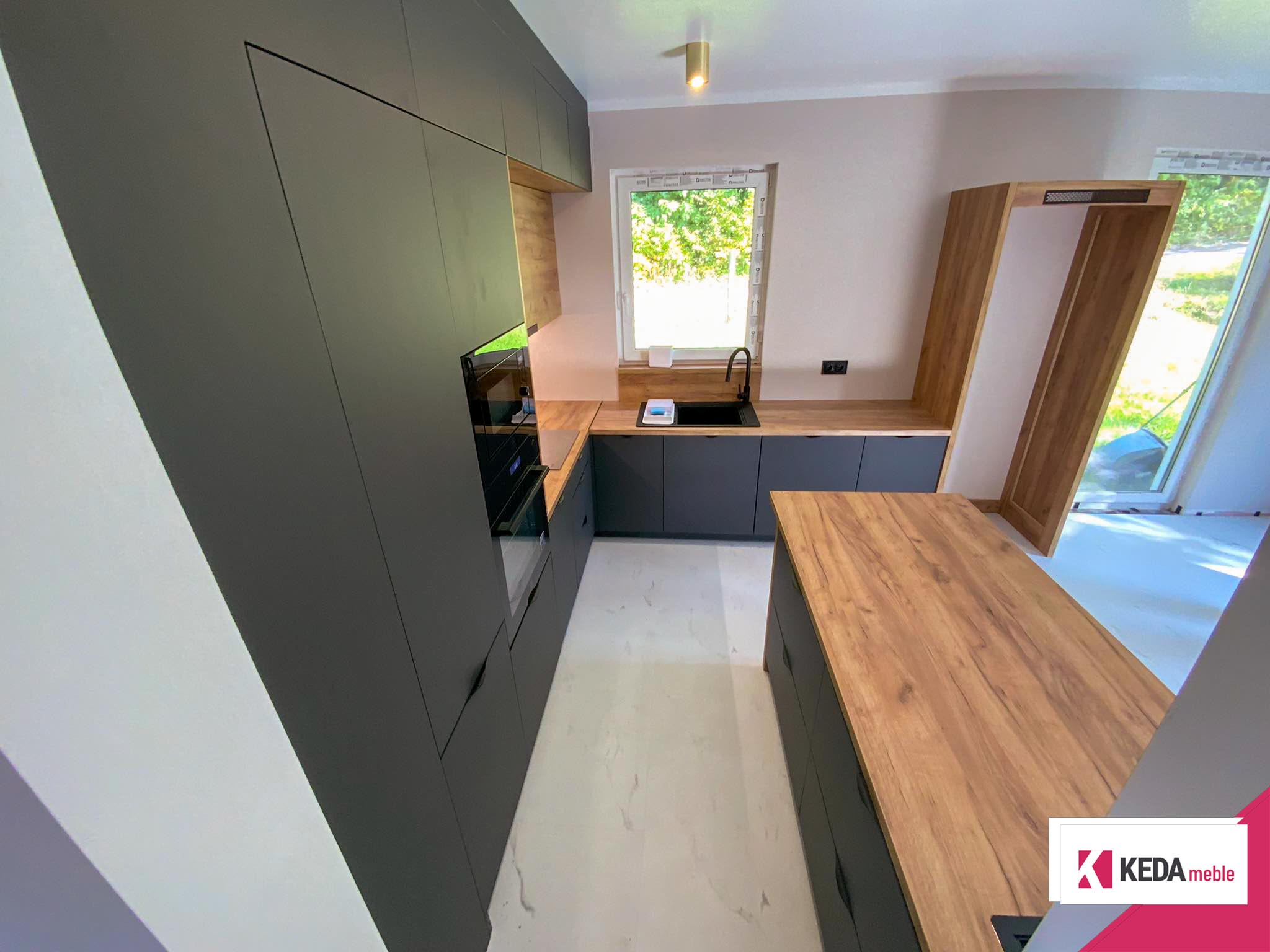 Nowoczesna kuchnia z szarymi szafkami i drewnianymi blatami, wbudowanymi urządzeniami AGD, dużym oknem z widokiem na zieleń oraz centralnie umieszczoną wyspą kuchenną.