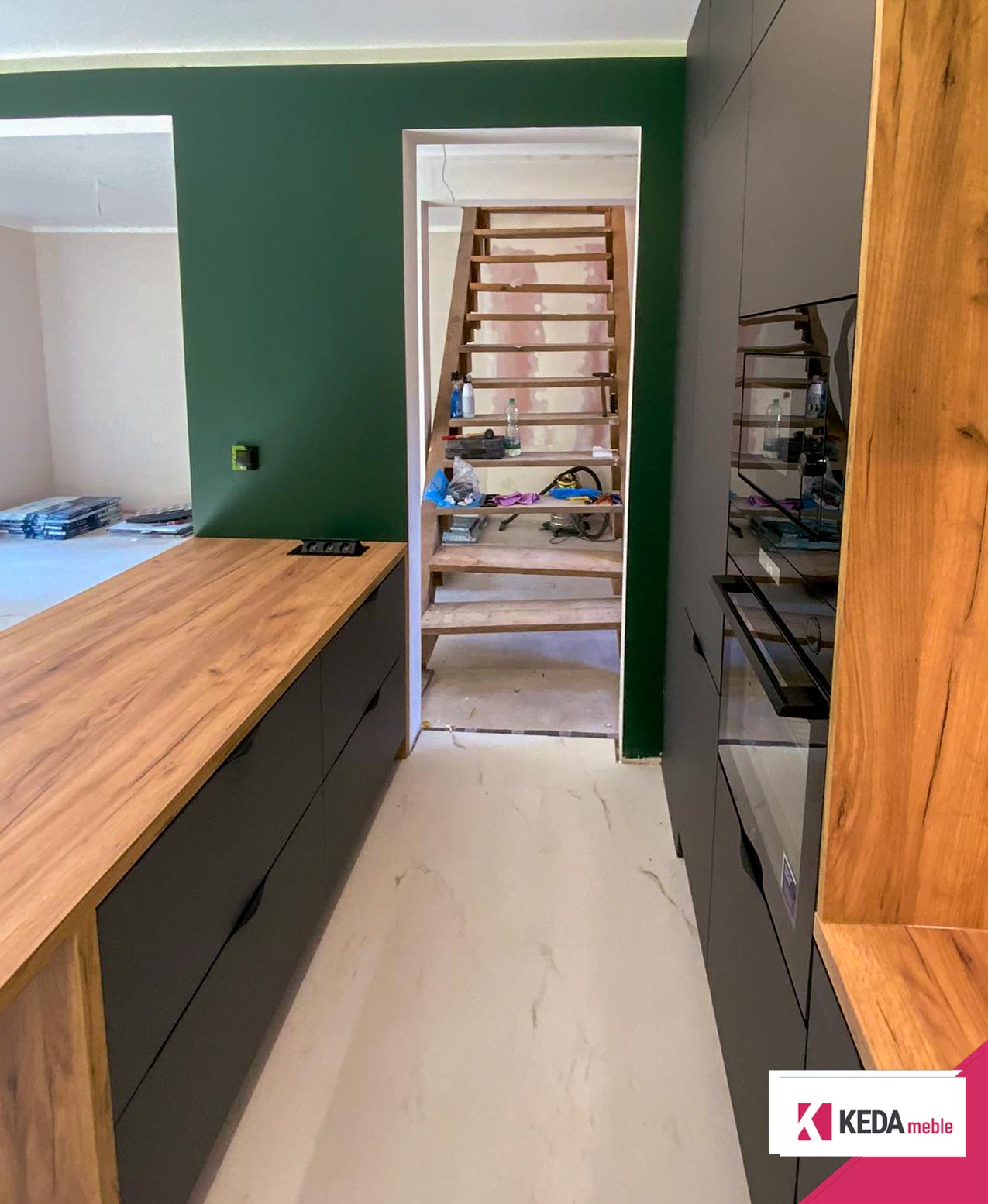 Nowoczesna kuchnia z szarymi szafkami i drewnianymi blatami, wbudowanymi urządzeniami AGD, dużym oknem z widokiem na zieleń oraz centralnie umieszczoną wyspą kuchenną.