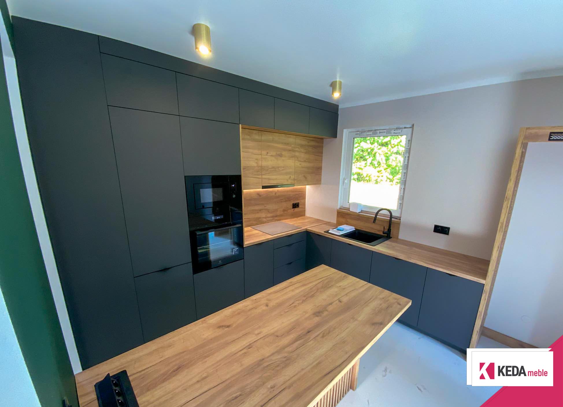 Nowoczesna kuchnia z szarymi szafkami i drewnianymi blatami, wbudowanymi urządzeniami AGD, dużym oknem z widokiem na zieleń oraz centralnie umieszczoną wyspą kuchenną.